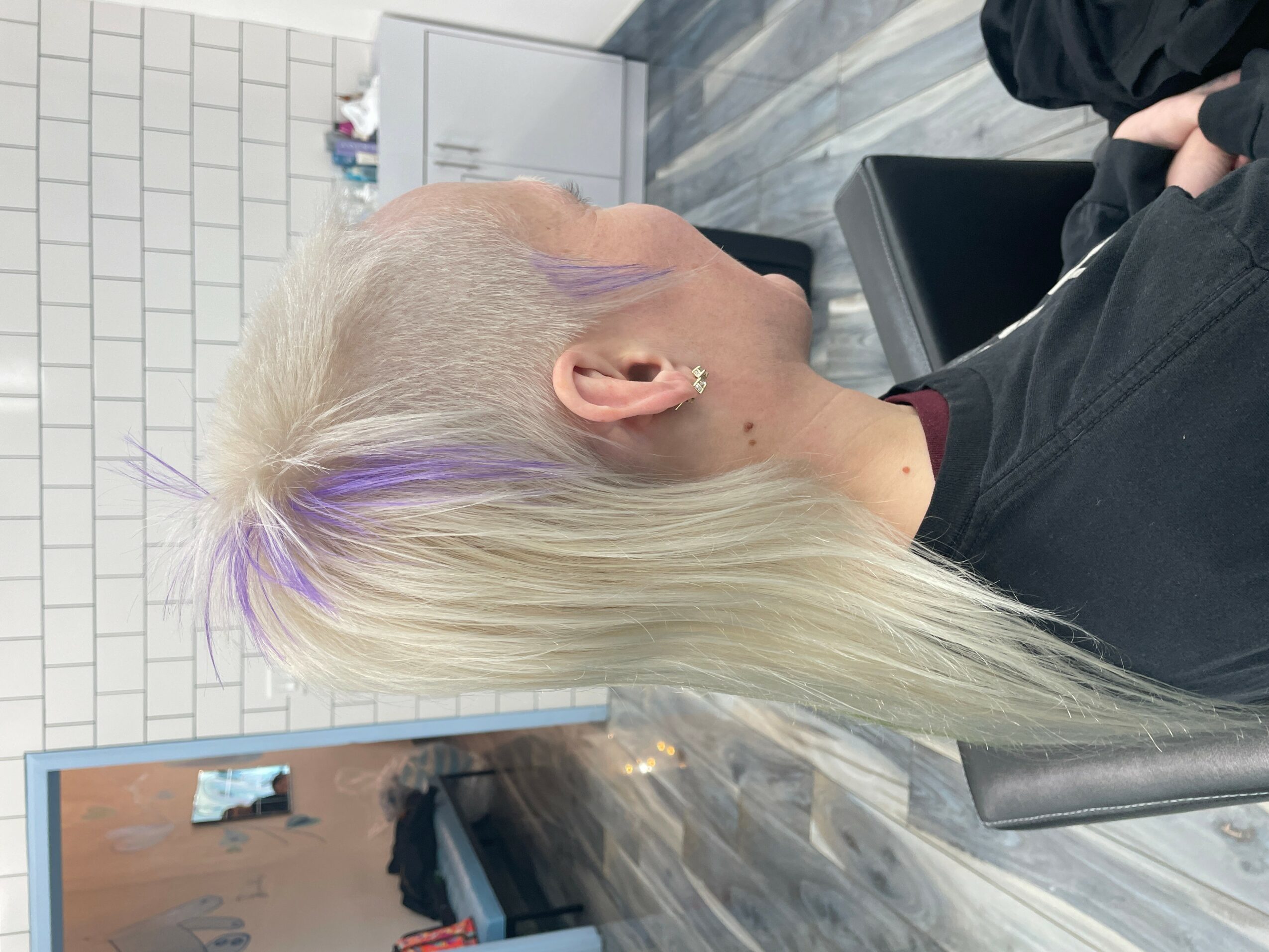 Person with platinum blonde hair and purple streaks, sitting in a salon chair, side view.