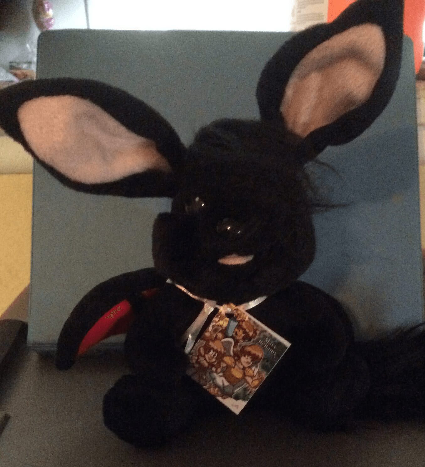Black plush bunny toy with large ears, wearing a gold chain and a colorful tag, sitting on a chair.