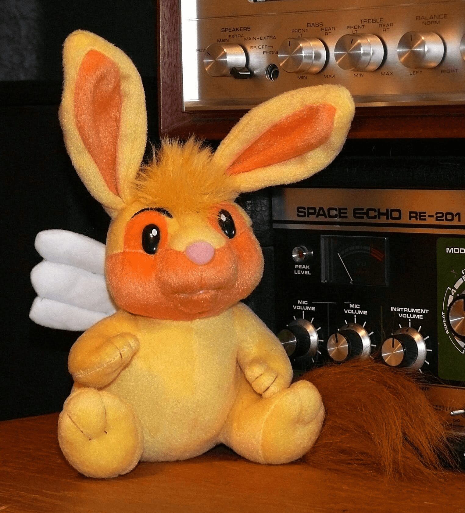 Plush bunny toy with large ears, orange face, pink nose, and white wings, sitting on a wooden surface with electronic equipment behind.
