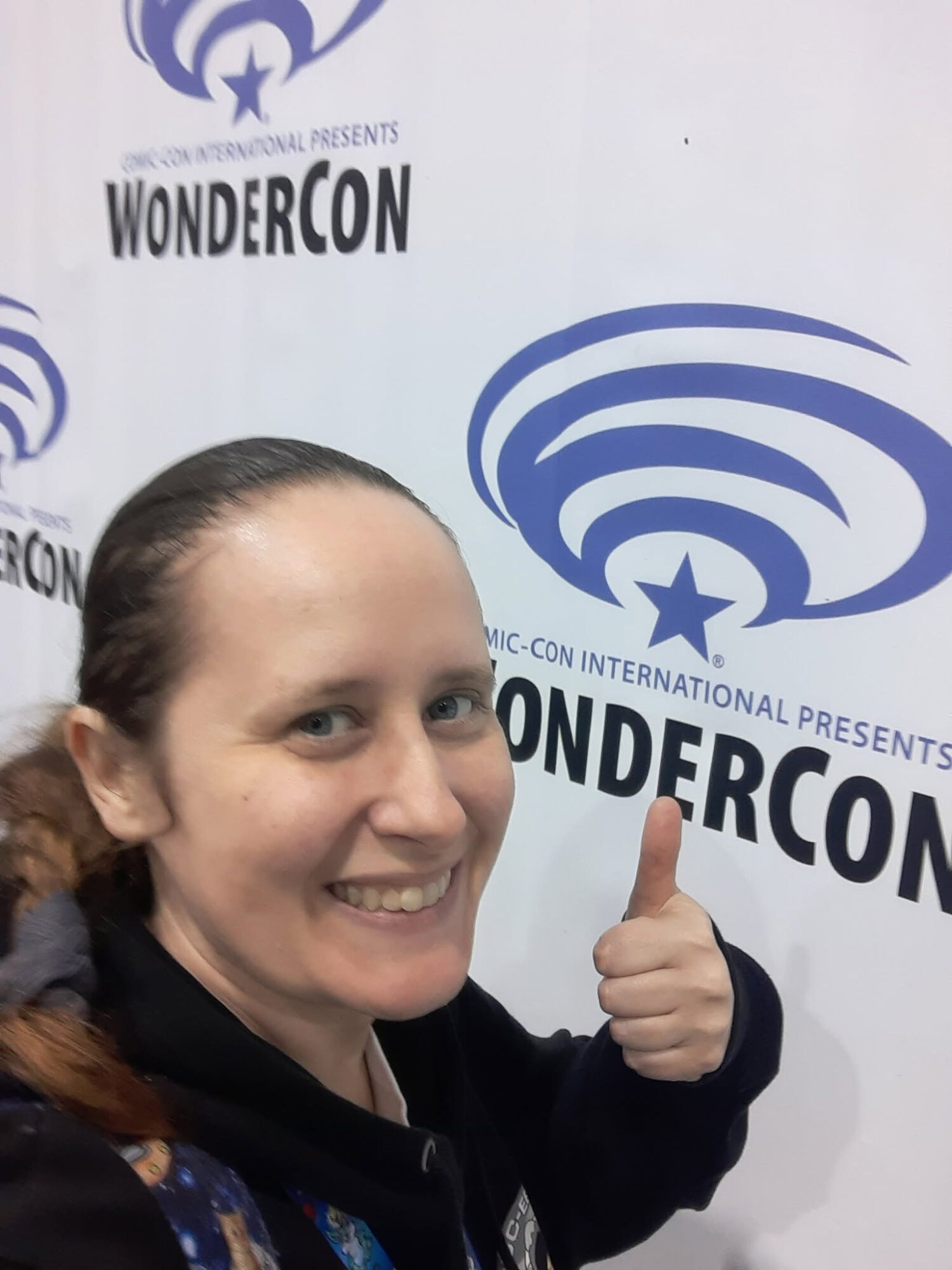 Woman smiling and giving a thumbs-up in front of WonderCon backdrop with logo and text.