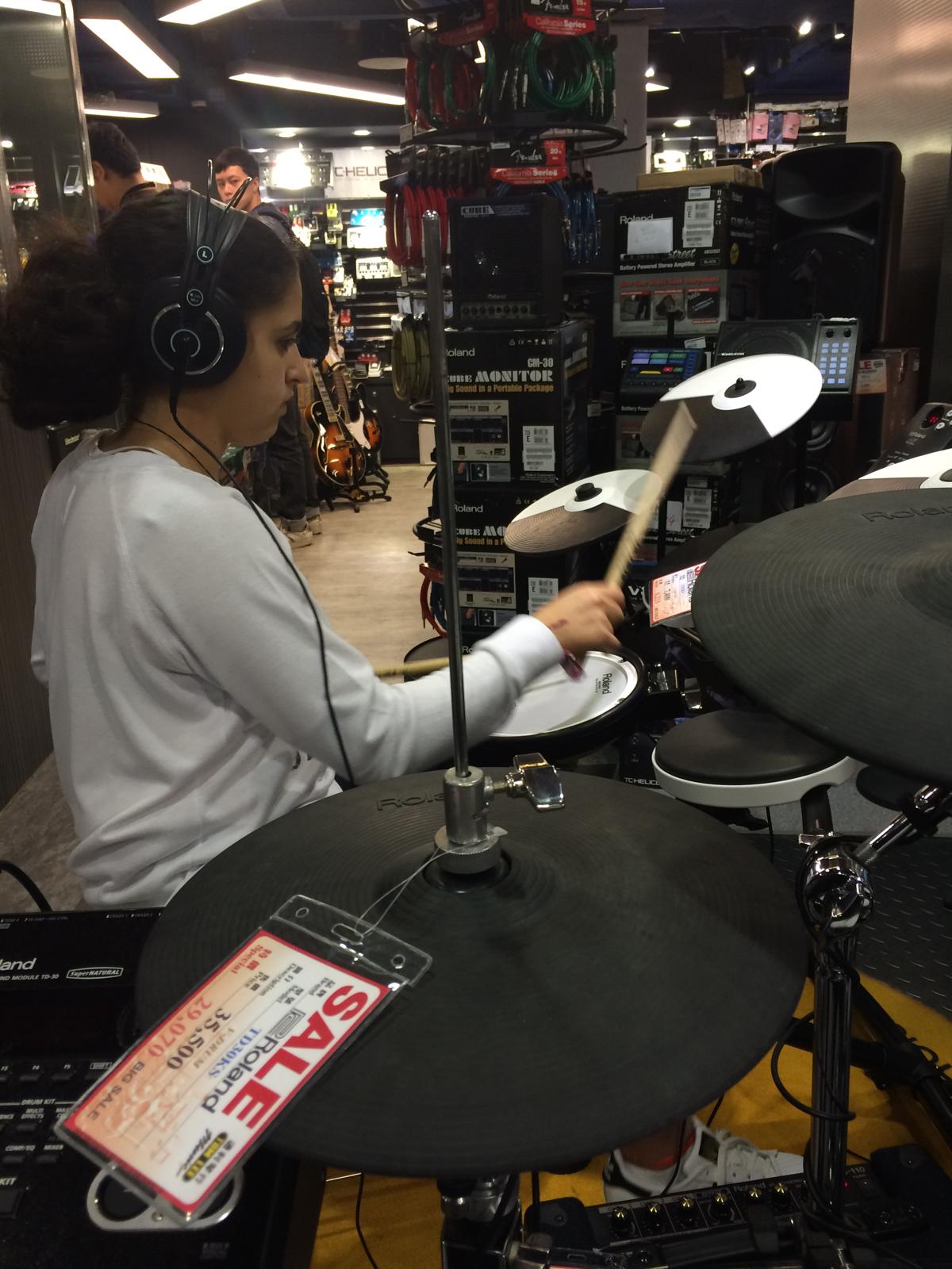 Young girl playing electronic drum set in a music store, wearing headphones, surrounded by musical equipment and displays.