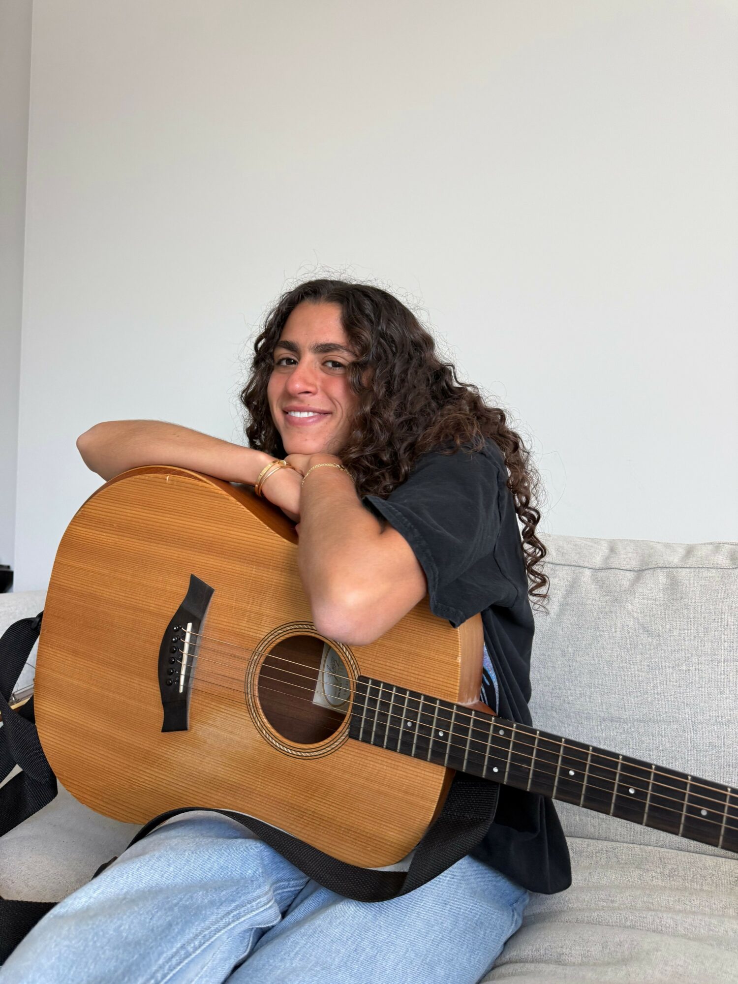 Person with curly hair smiling, hugging an acoustic guitar, sitting on a light-colored couch, wearing a black shirt and blue jeans.