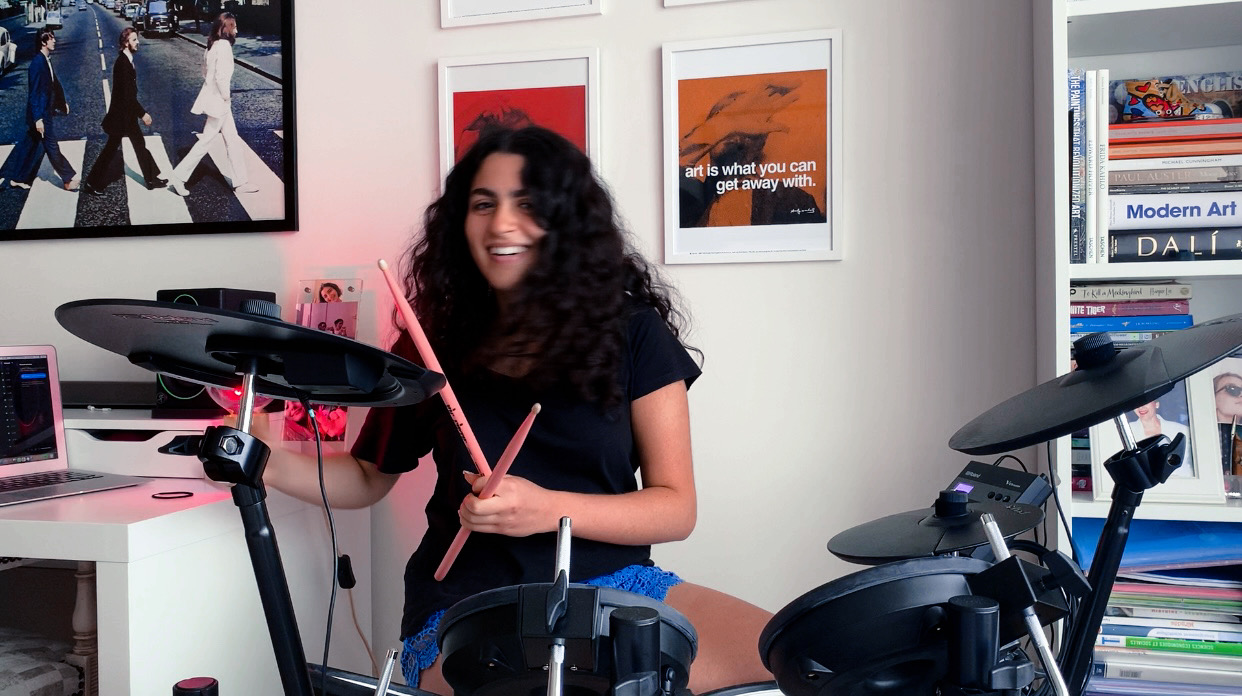 Young woman with curly hair smiling in a room with artwork and books, surrounded by electronic drum pads and a laptop.