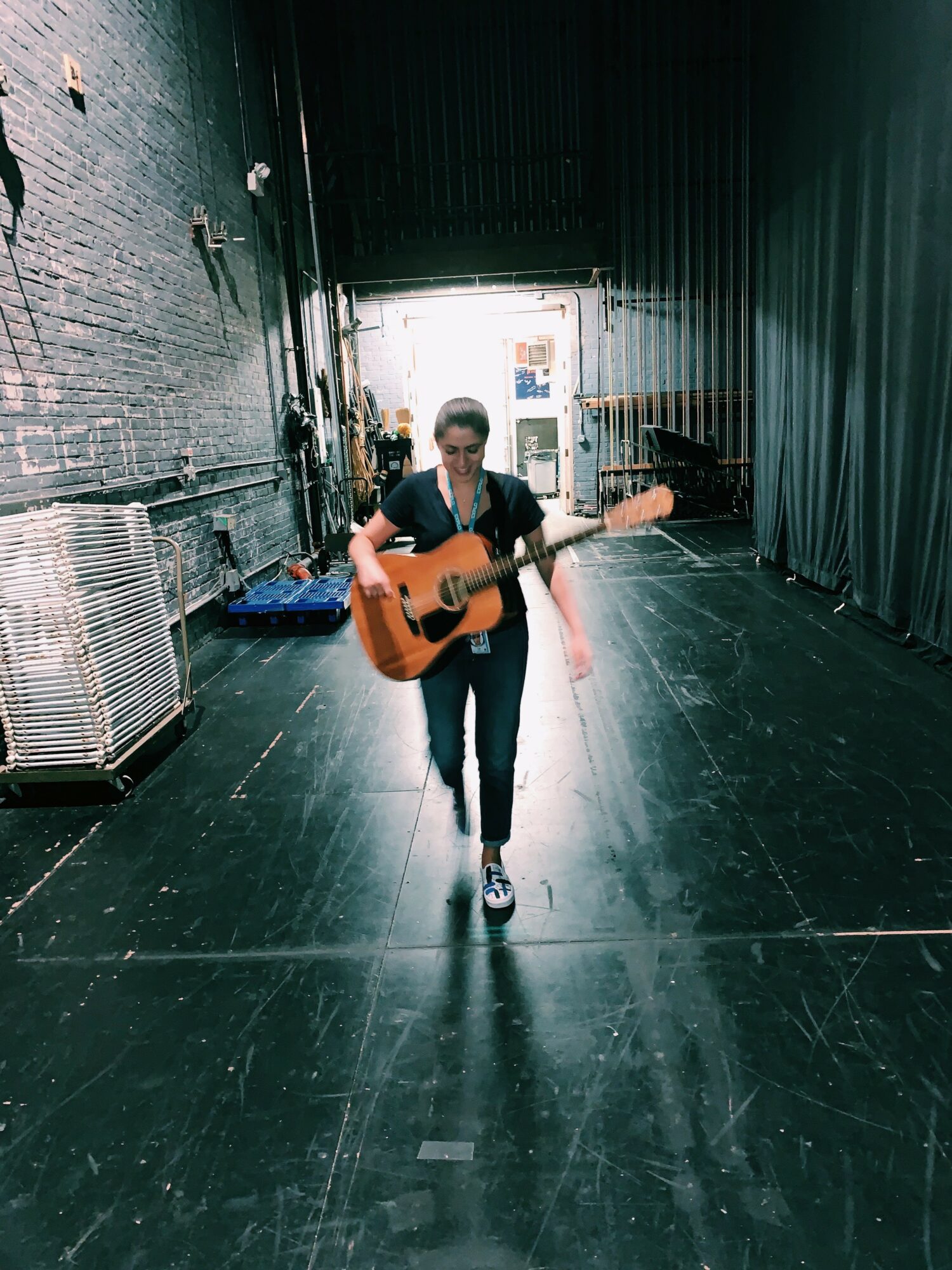 Person walking with guitar in a dark backstage area, bright light at the exit, industrial walls, and equipment on the sides.