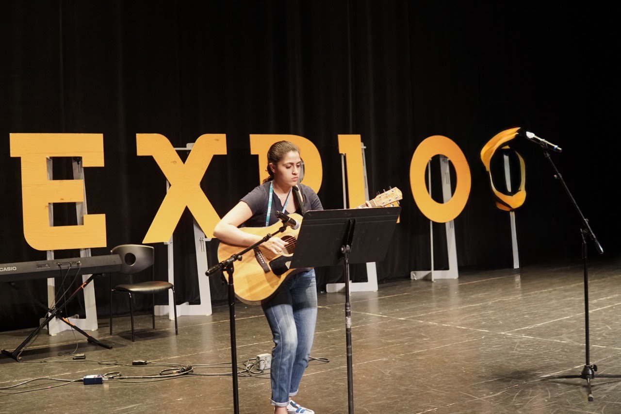 Young woman playing guitar on stage with large yellow letters spelling 'EXPO' behind her, microphone stand nearby.