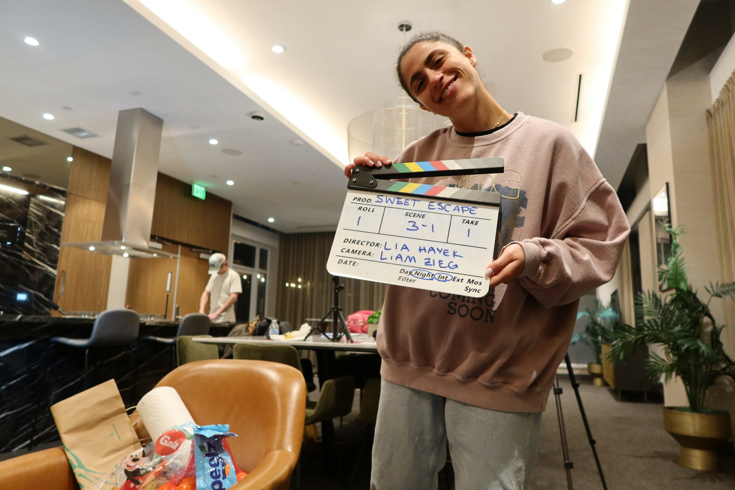 Woman holding a clapperboard in a modern indoor space with tables, chairs, and plants.