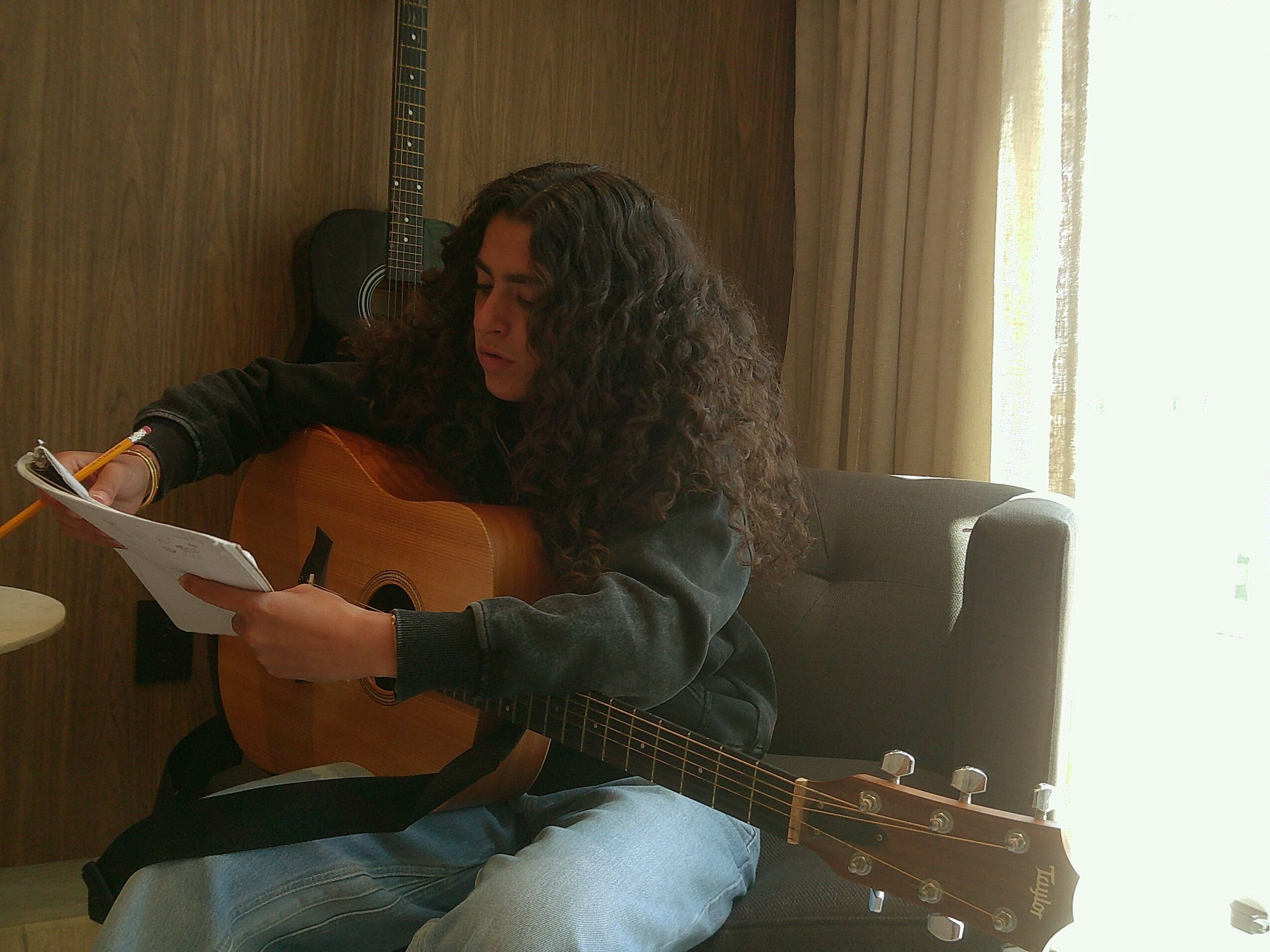 Person with curly hair sitting on a chair, playing an acoustic guitar and reading a paper, with a guitar on the wall behind.