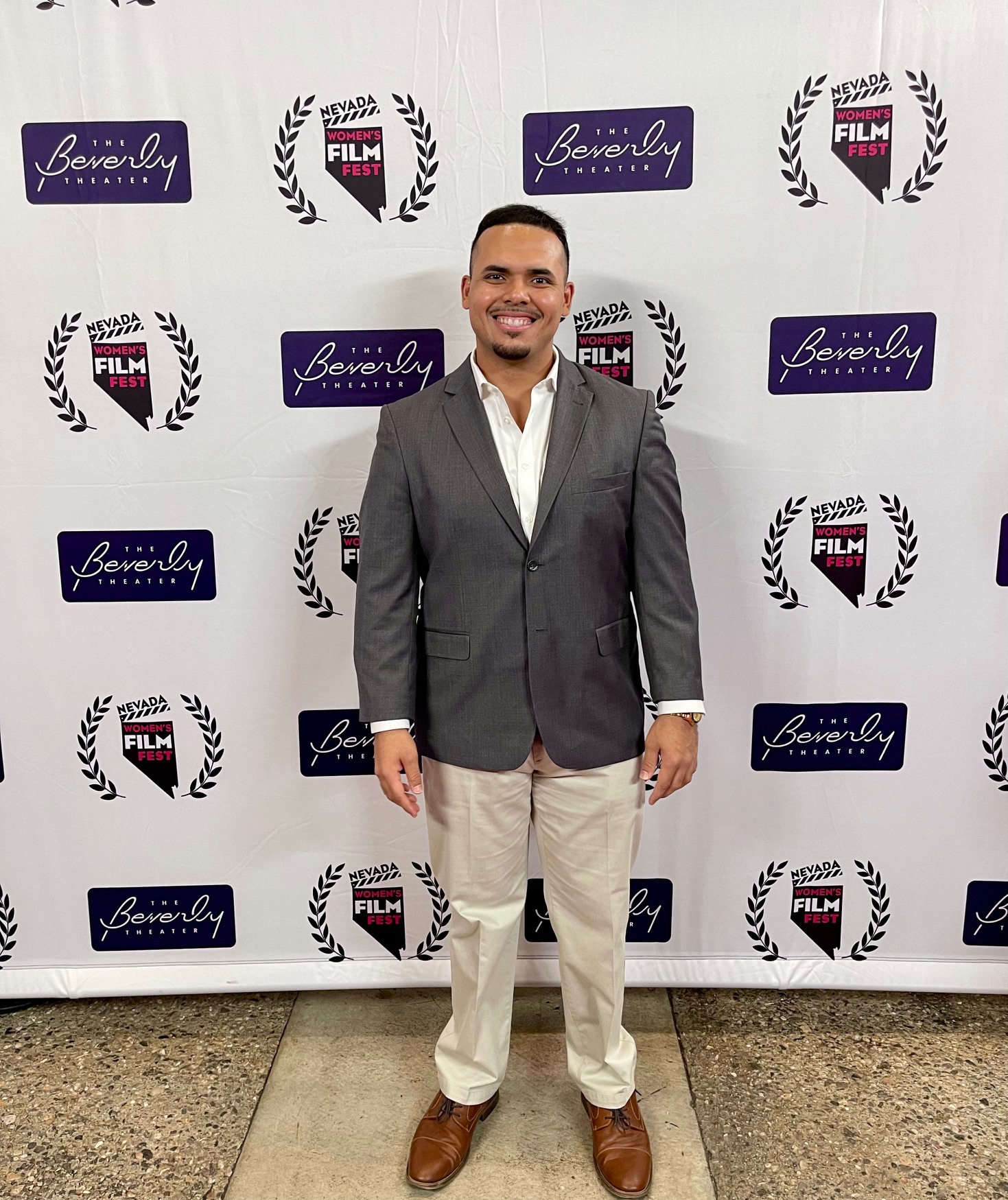Man in a gray blazer, white shirt, and beige pants standing in front of a backdrop with logos, smiling.