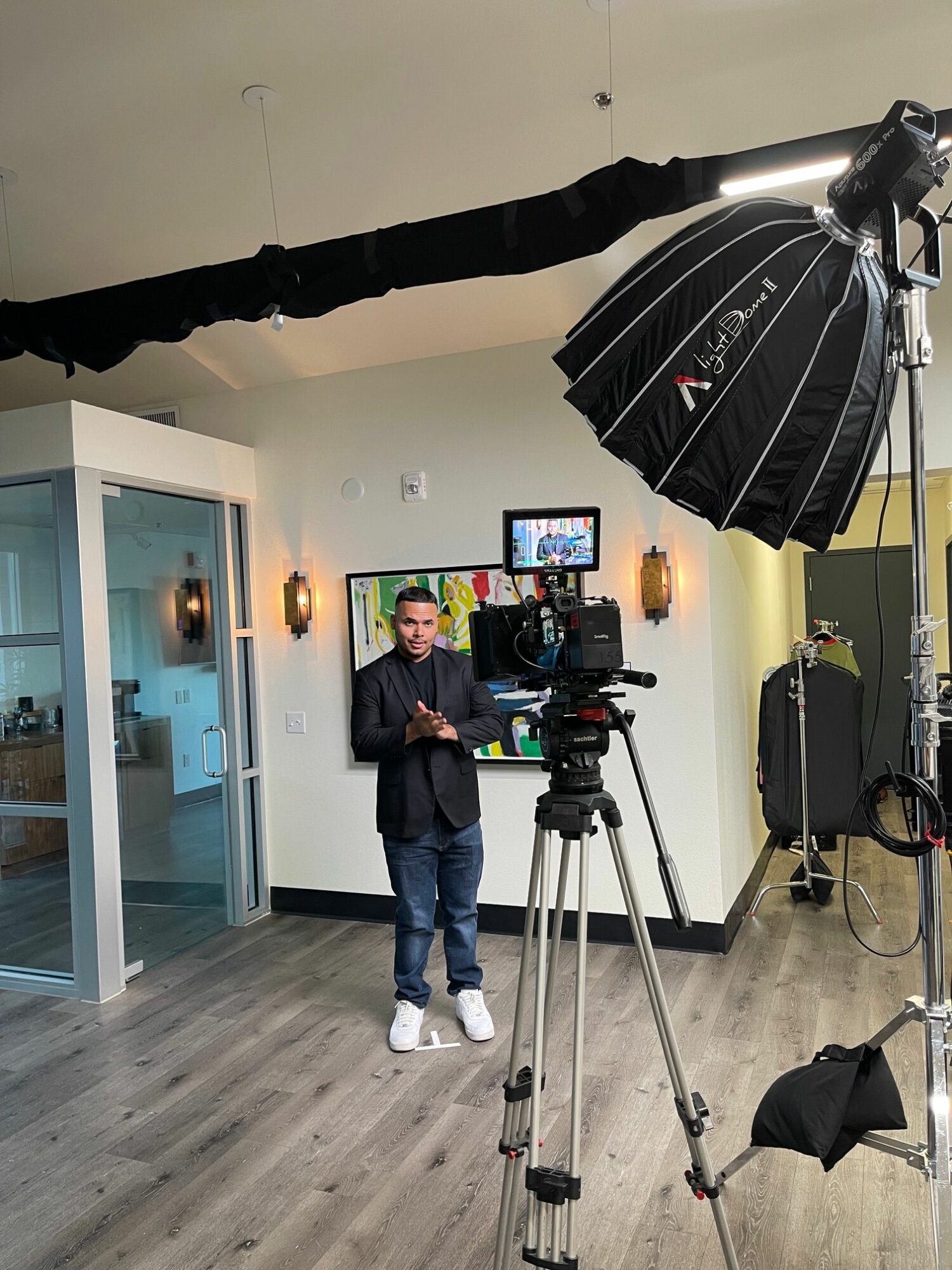 Photographer in a studio with camera equipment, standing in front of a TV screen, with a person in the background, and studio lights.