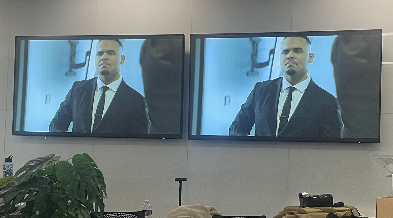 Two screens display a man in a suit and tie, sitting indoors with a plant in the foreground.