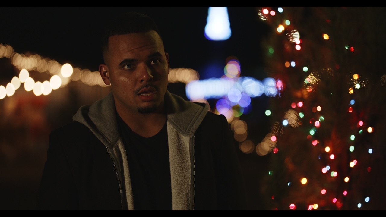 A man stands outdoors at night with blurred colorful lights in the background.