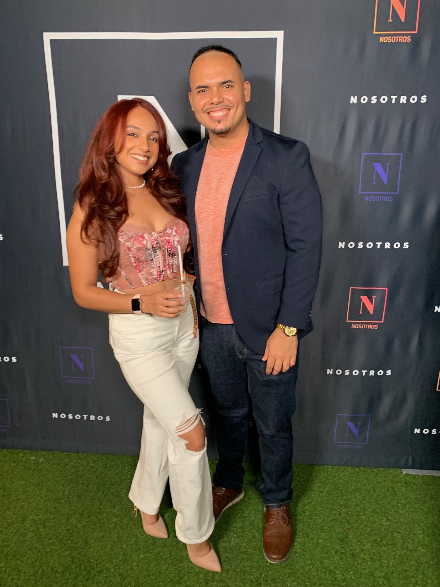A woman with long red hair and a man with short dark hair stand together, smiling, in front of a backdrop with logos.