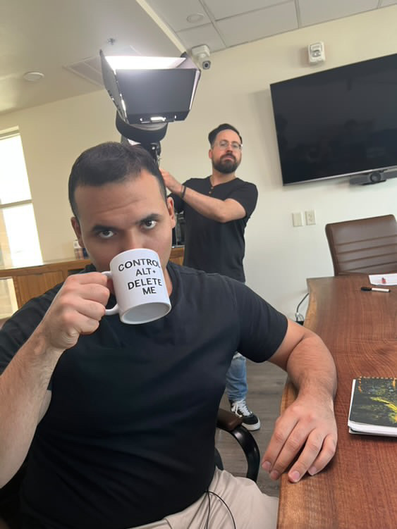 Man with short dark hair drinking from a mug, sitting at a table in a room with a TV and a person in the background.