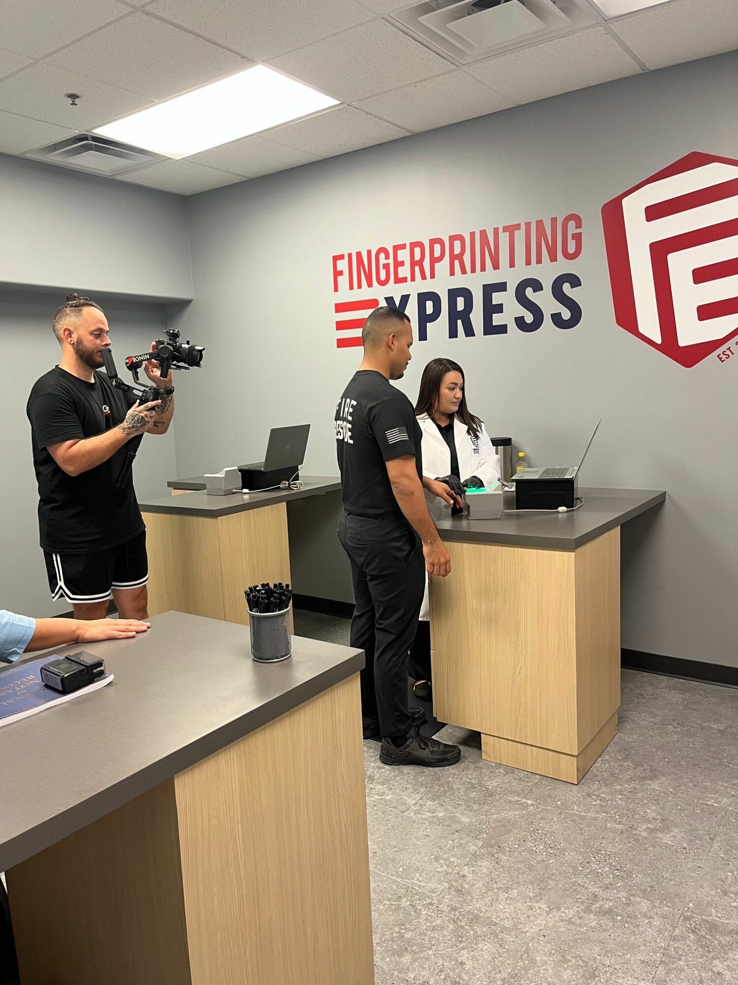 Person taking a selfie in an office with a sign reading 'FINGERPRINTING EXPRESS' on the wall, holding a phone, with a camera operator nearby.