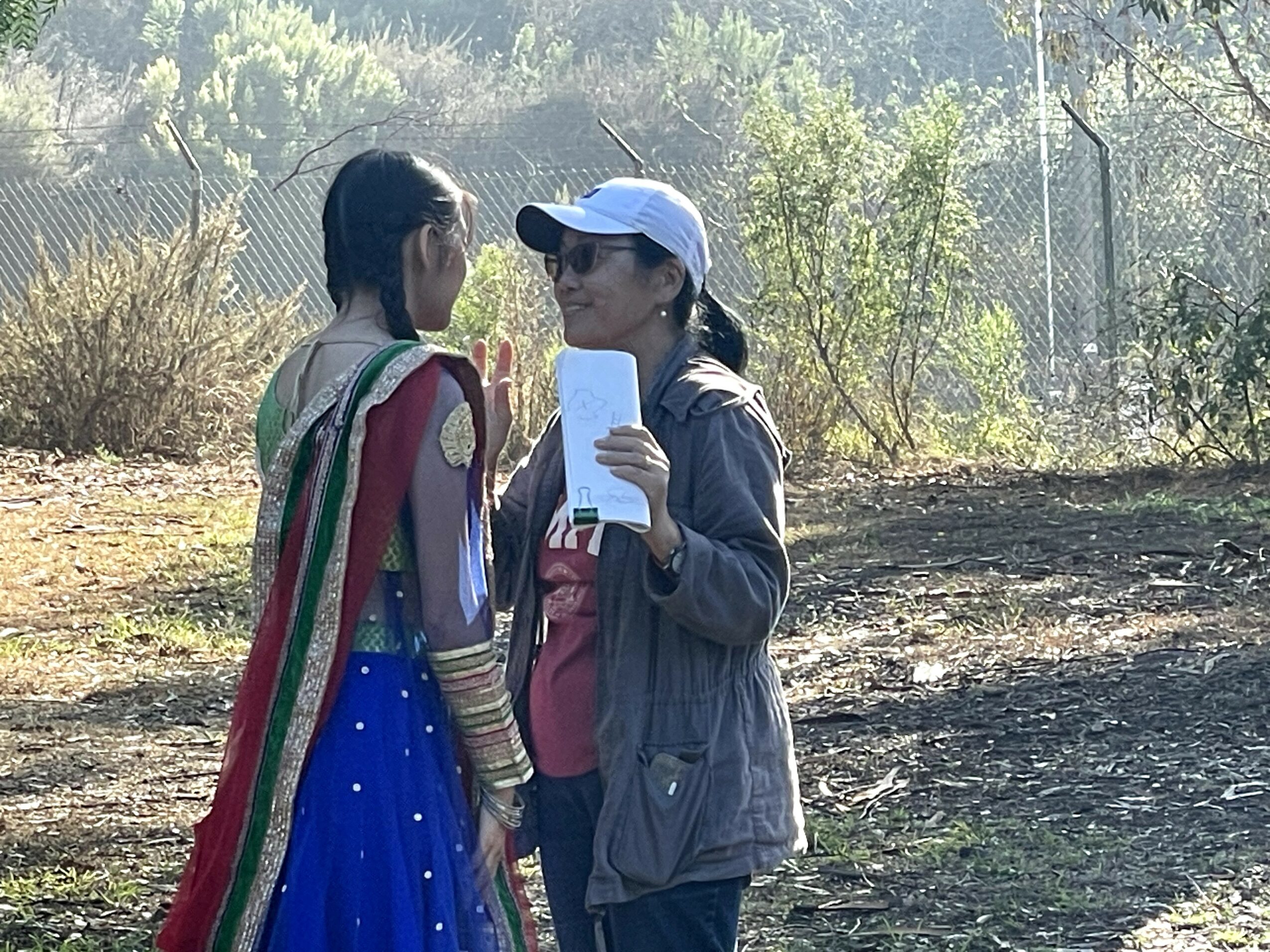 Two women stand outdoors, talking, with trees and a fence in the background.