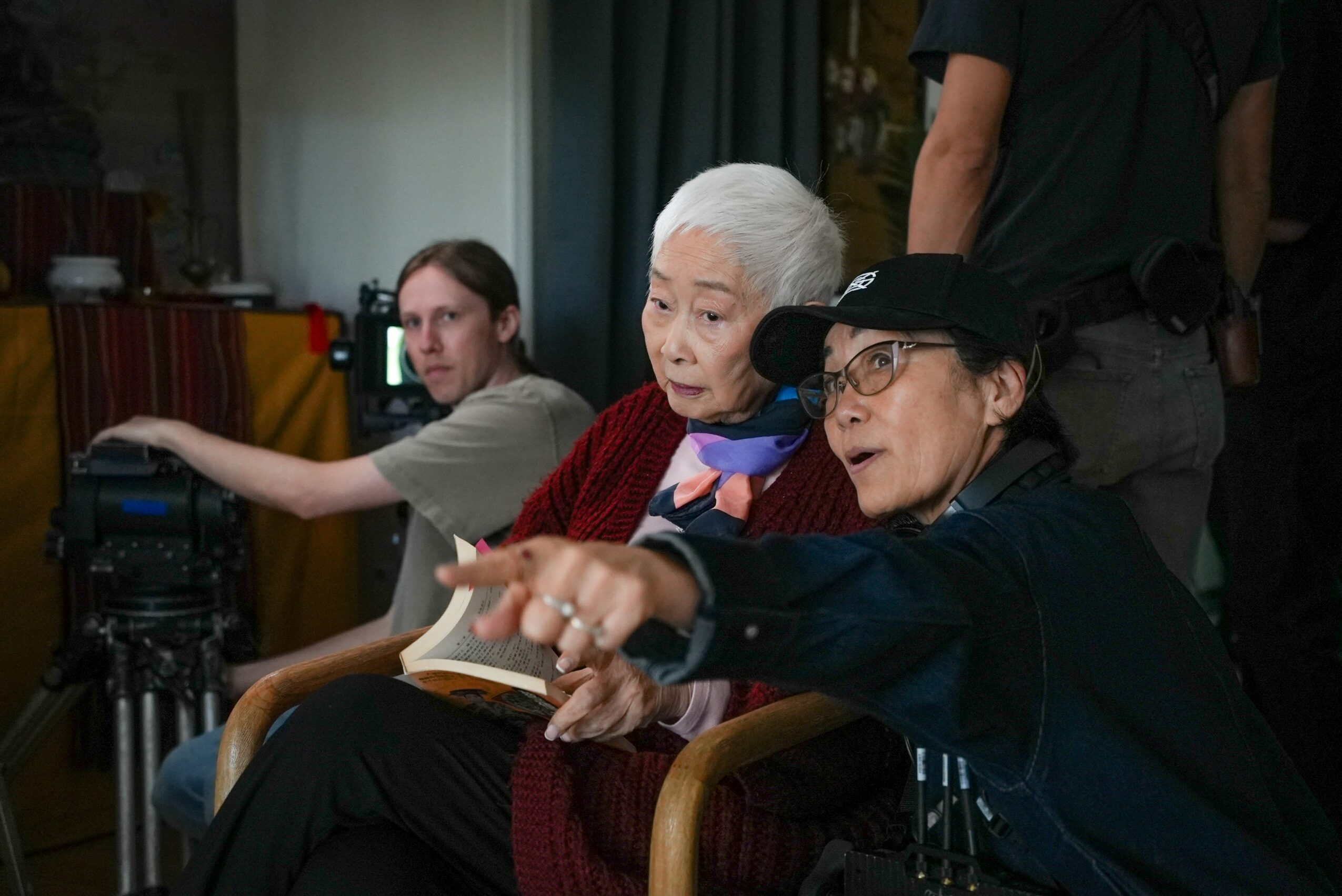 Two women sit and look at a screen, one pointing, with a person operating a camera nearby.