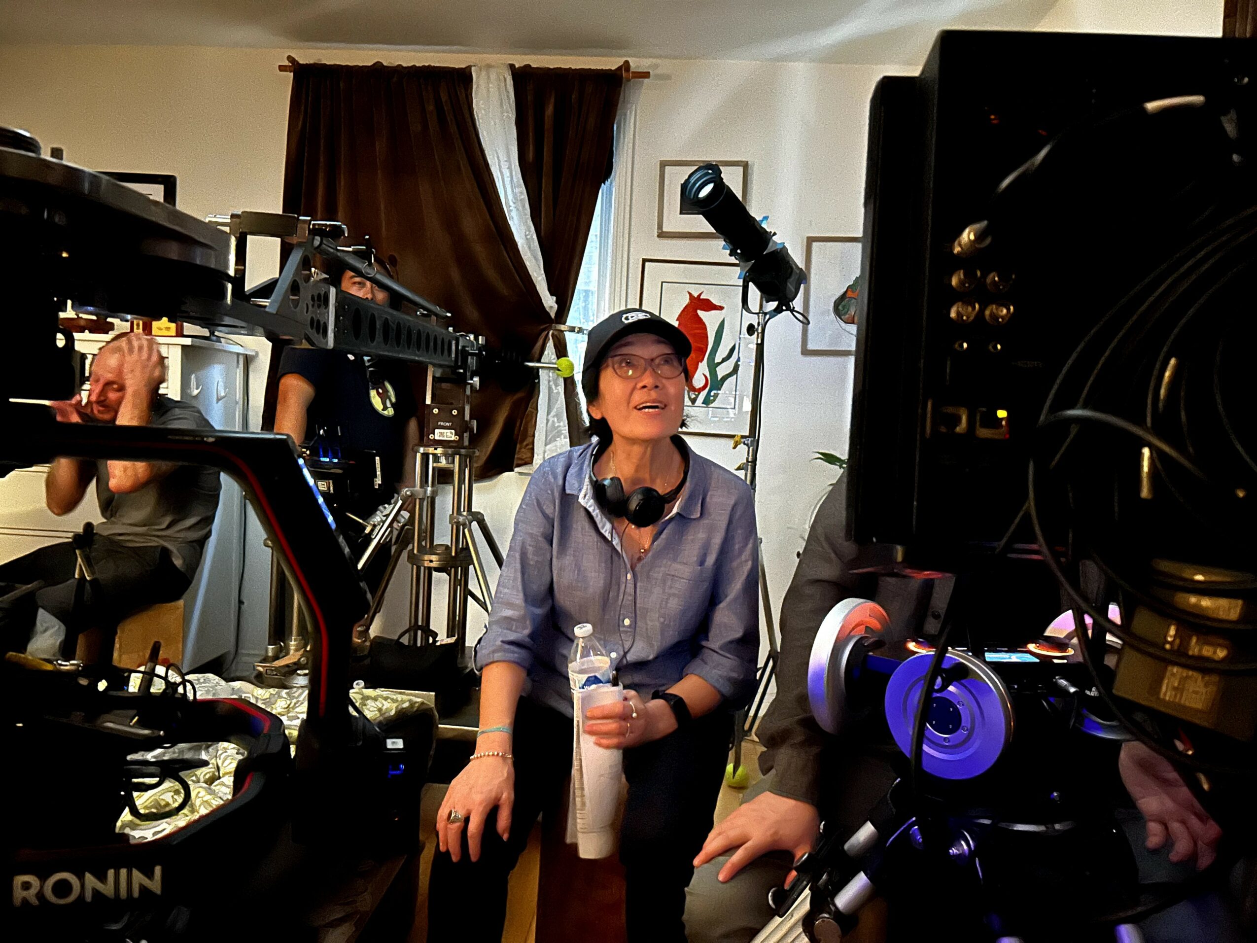 Person with glasses and headphones in a recording studio, surrounded by cameras and equipment, holding a drink and smiling.