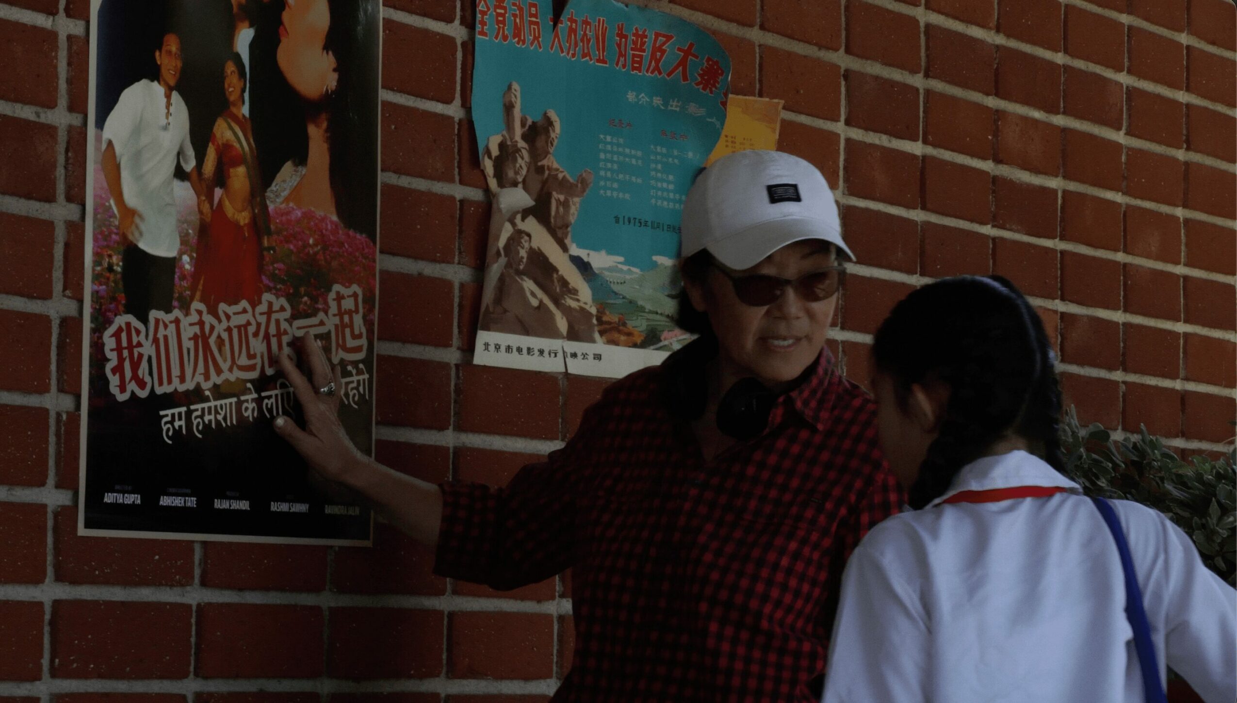 Two people talking in front of a brick wall with posters, one wearing a white cap and glasses, the other with long hair.