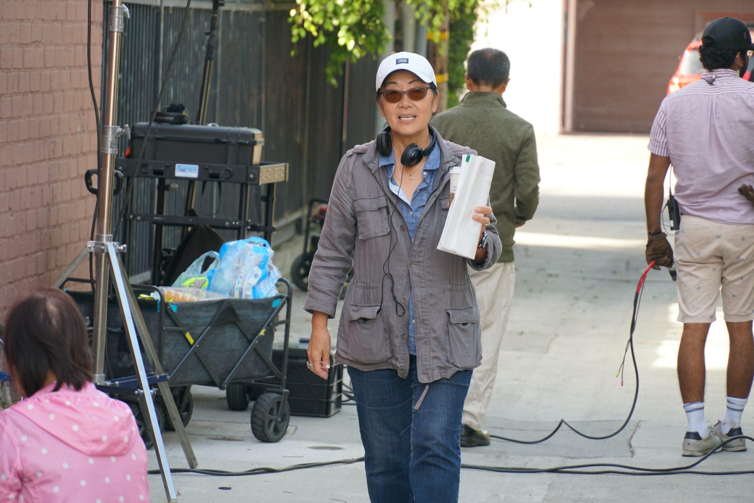 Woman wearing sunglasses, a cap, and a gray jacket holding papers, walking outdoors with people in the background.