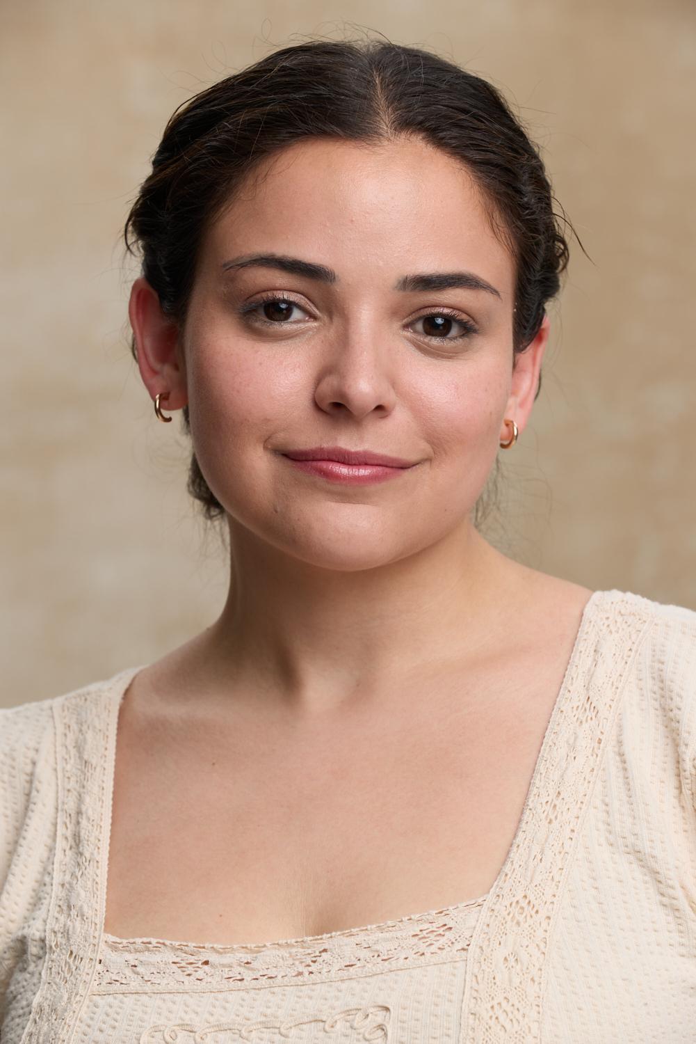 Young woman with dark hair pulled back, wearing a cream-colored top, smiling softly, neutral background.