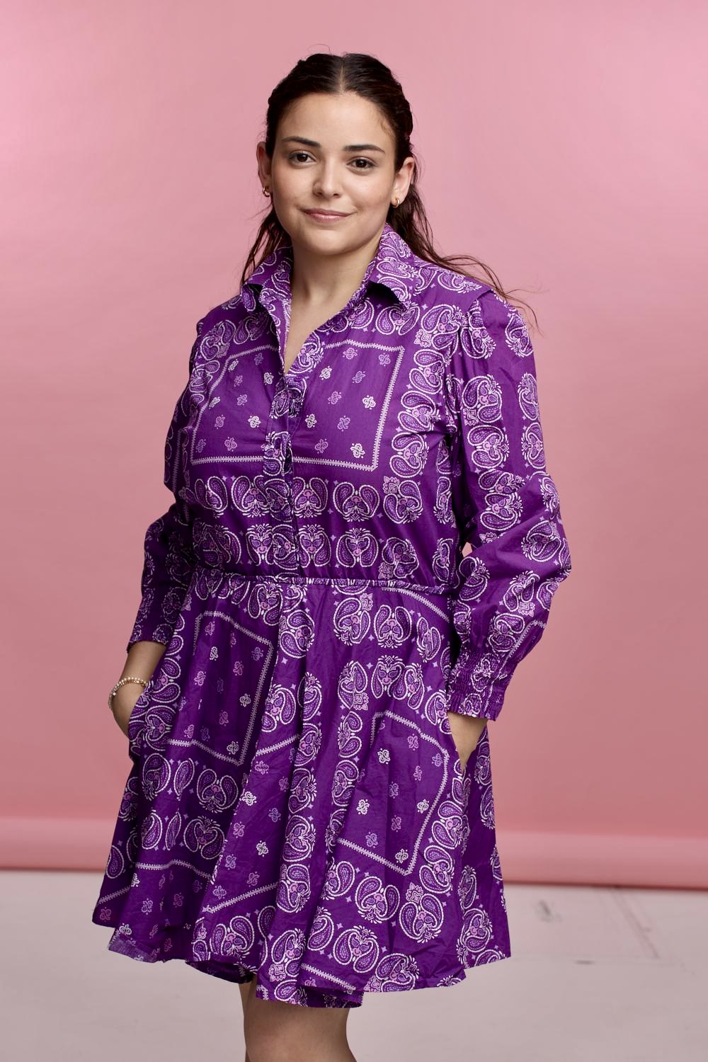 Young woman with long hair wearing a purple patterned dress, standing against a pink background.