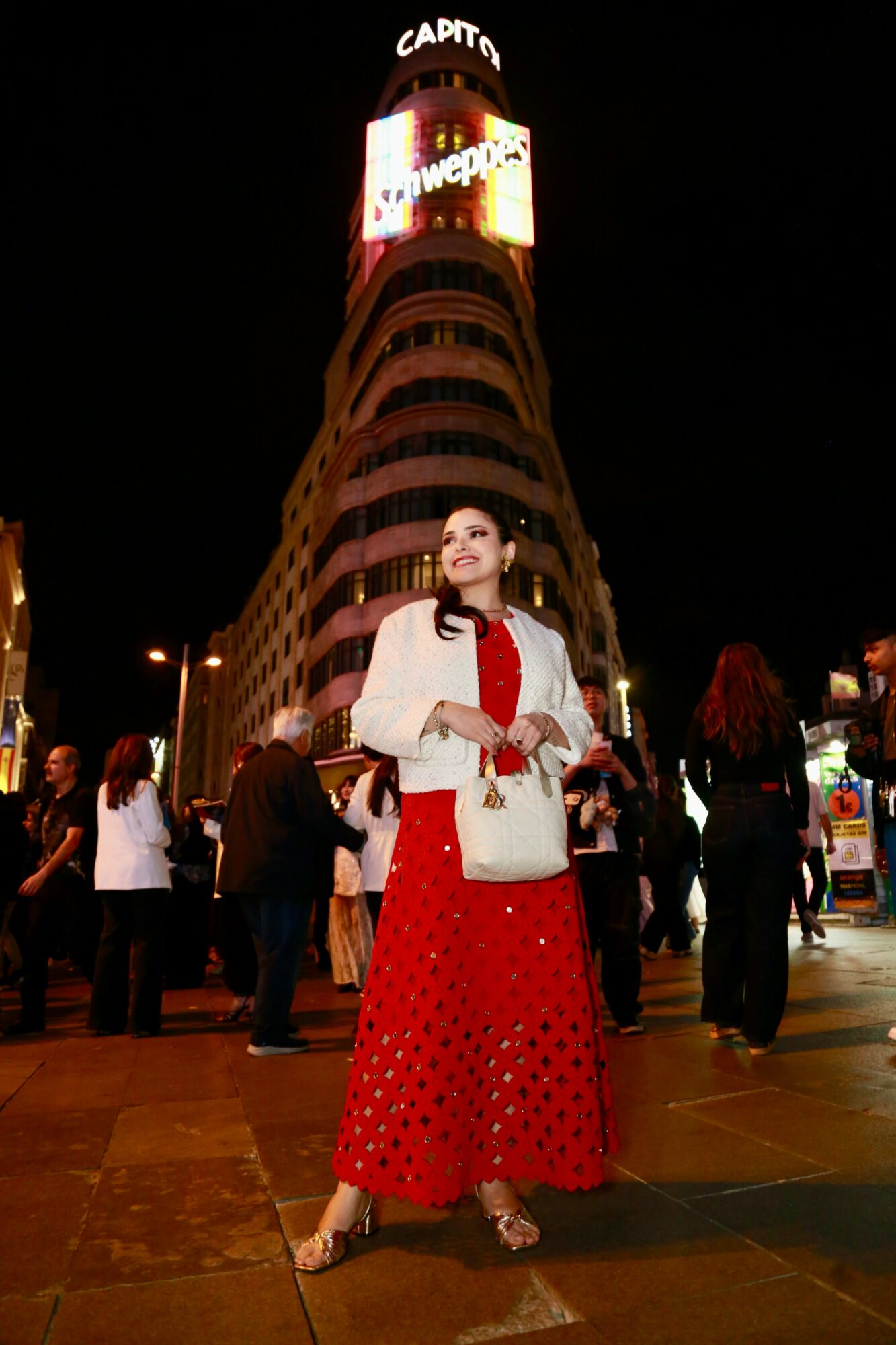 Woman in red dress and white jacket standing in city square at night with tall building and illuminated signs behind her.