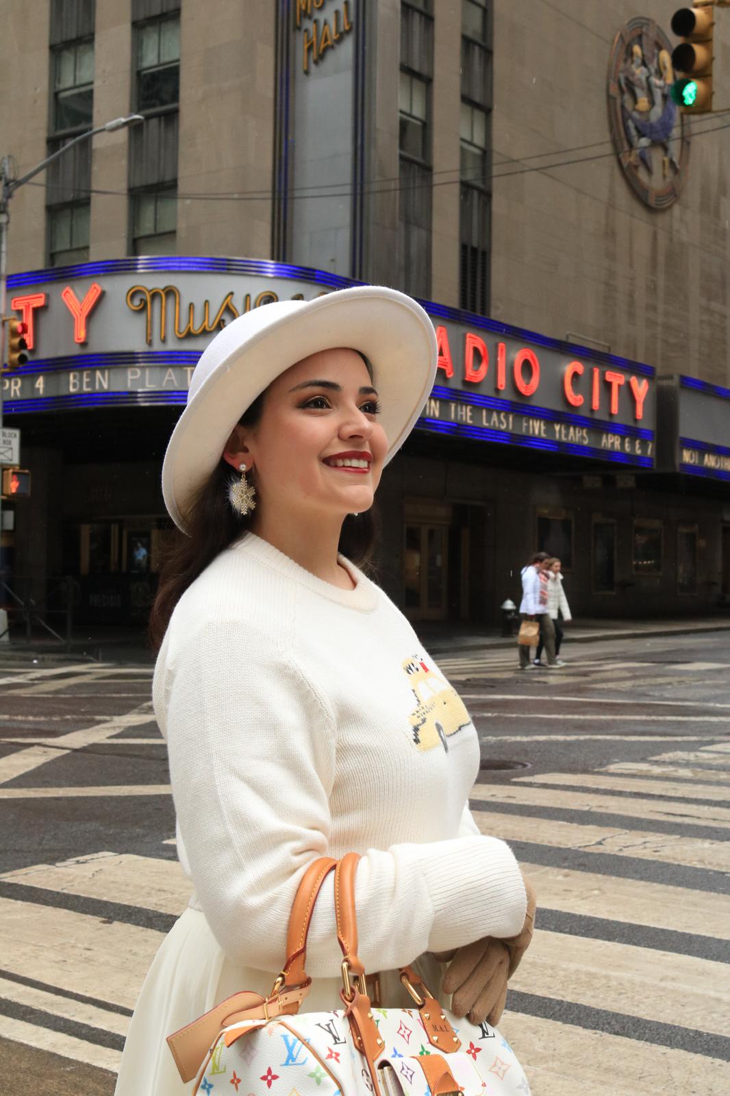 Smiling woman in white hat and sweater holding a handbag on city street with theater marquee in background.