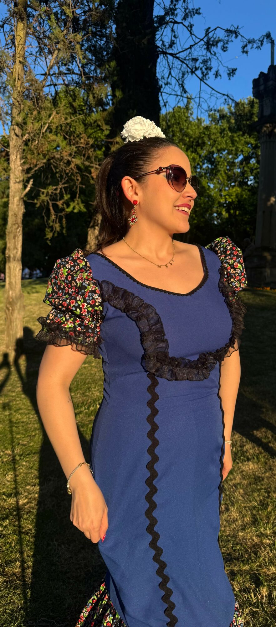 Woman wearing sunglasses and a blue dress with puffed sleeves, standing outdoors with trees and blue sky in background.