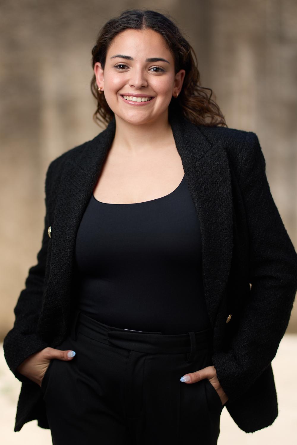 Smiling woman with dark hair, wearing a black blazer and top, standing outdoors with a blurred background.