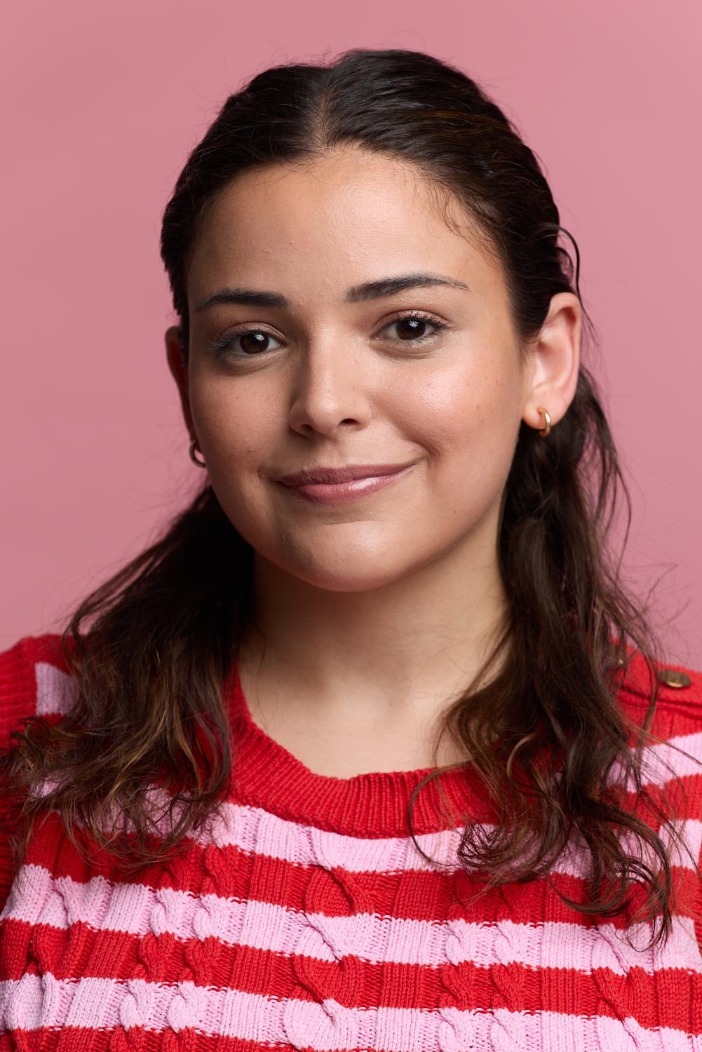 Young woman with dark hair and a red and white striped sweater, smiling against a pink background.