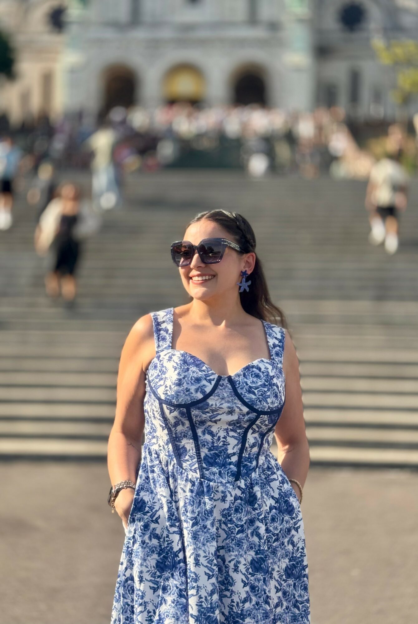 Woman in sunglasses and a blue and white patterned dress standing outdoors on steps, smiling, with people in the background.