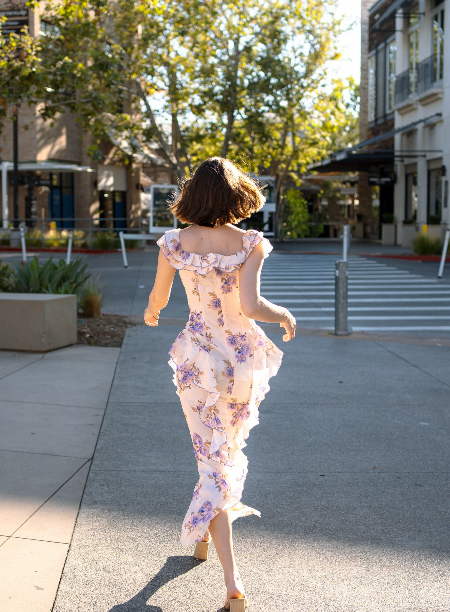 Young girl with short hair walking on sidewalk in sunlight, wearing floral dress, trees, buildings, and crosswalk in background.