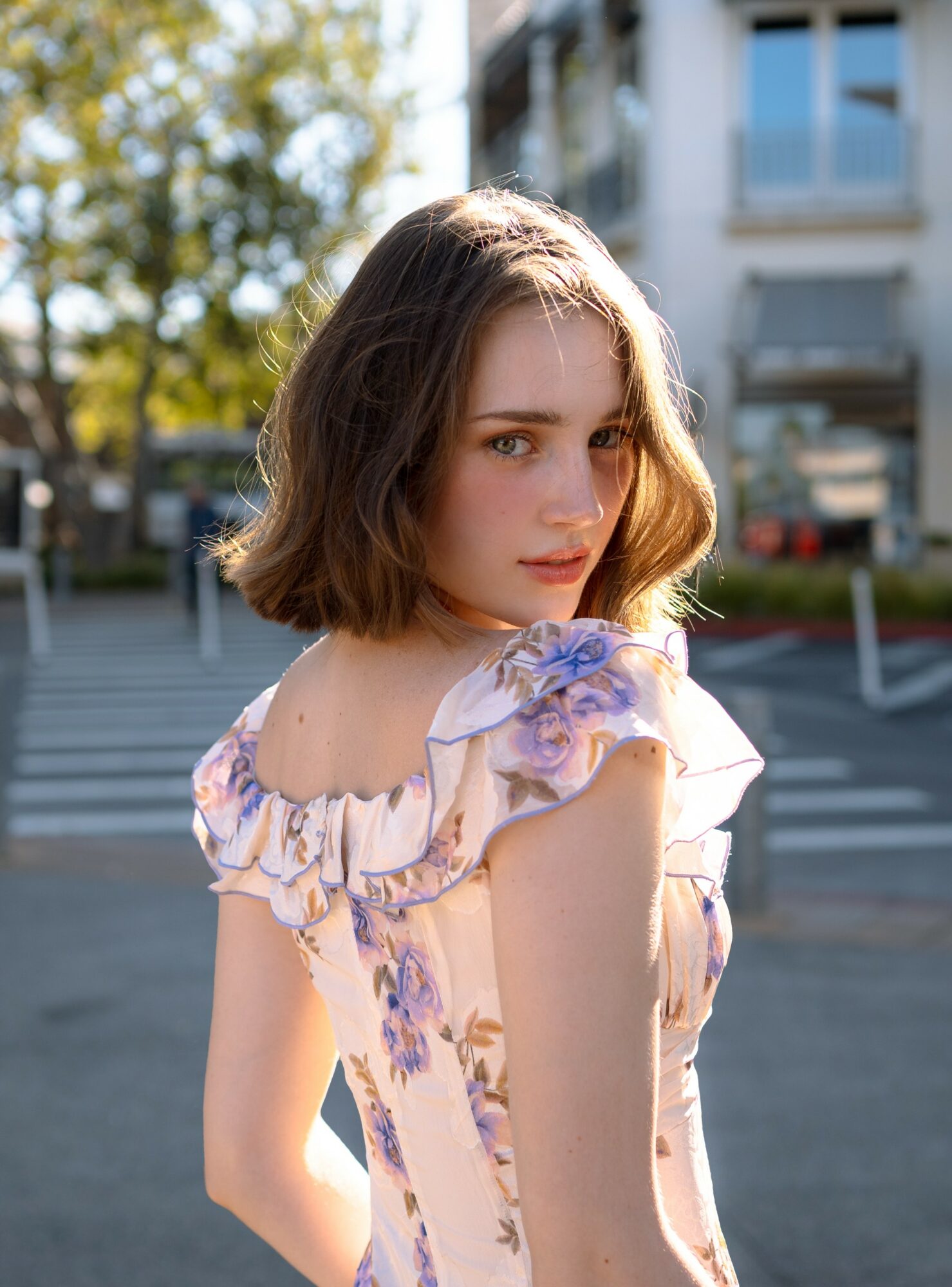 Young woman with shoulder-length brown hair in a floral dress outdoors, sunlight, urban background