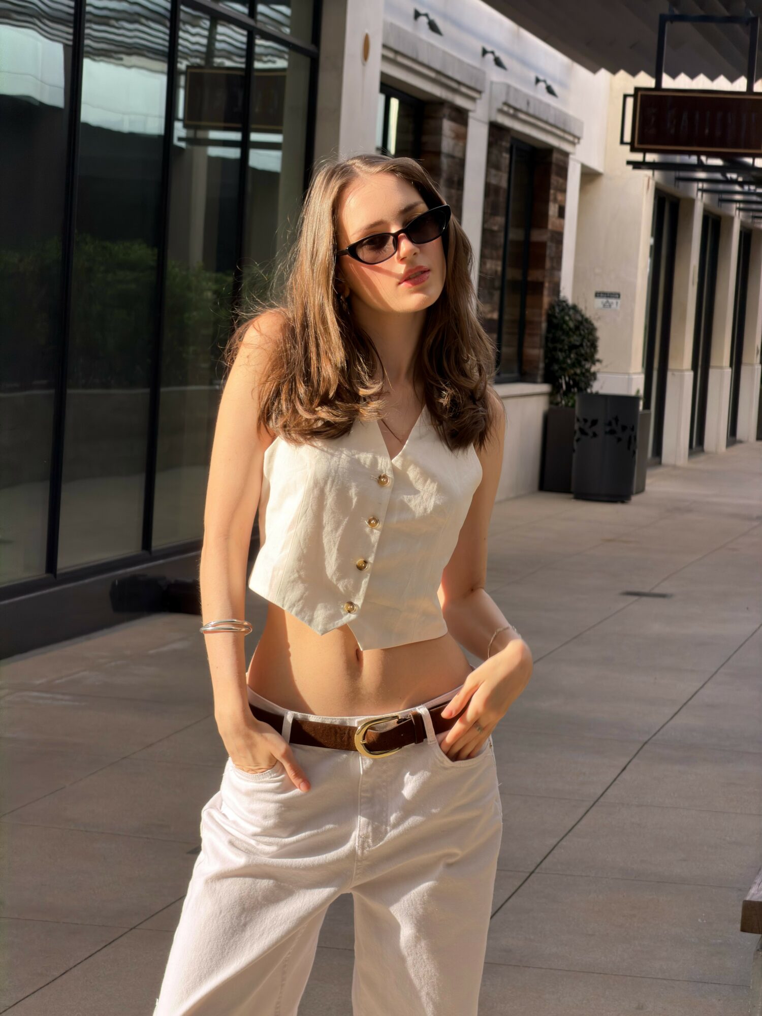 Young woman with long hair wearing sunglasses, a sleeveless top, and white pants, standing outdoors in sunlight.