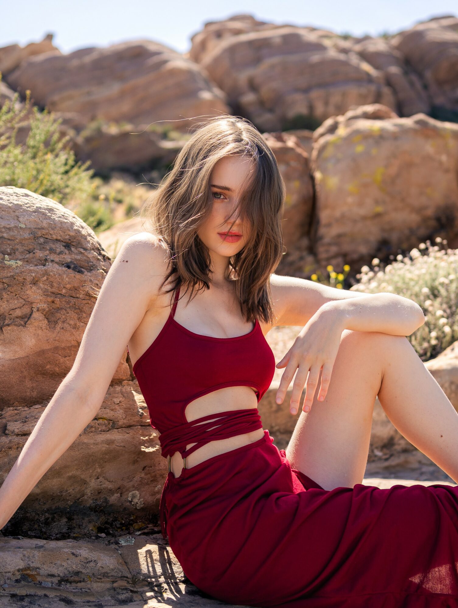 Young woman with shoulder-length hair in a red dress sitting on rocks outdoors, with desert-like background.