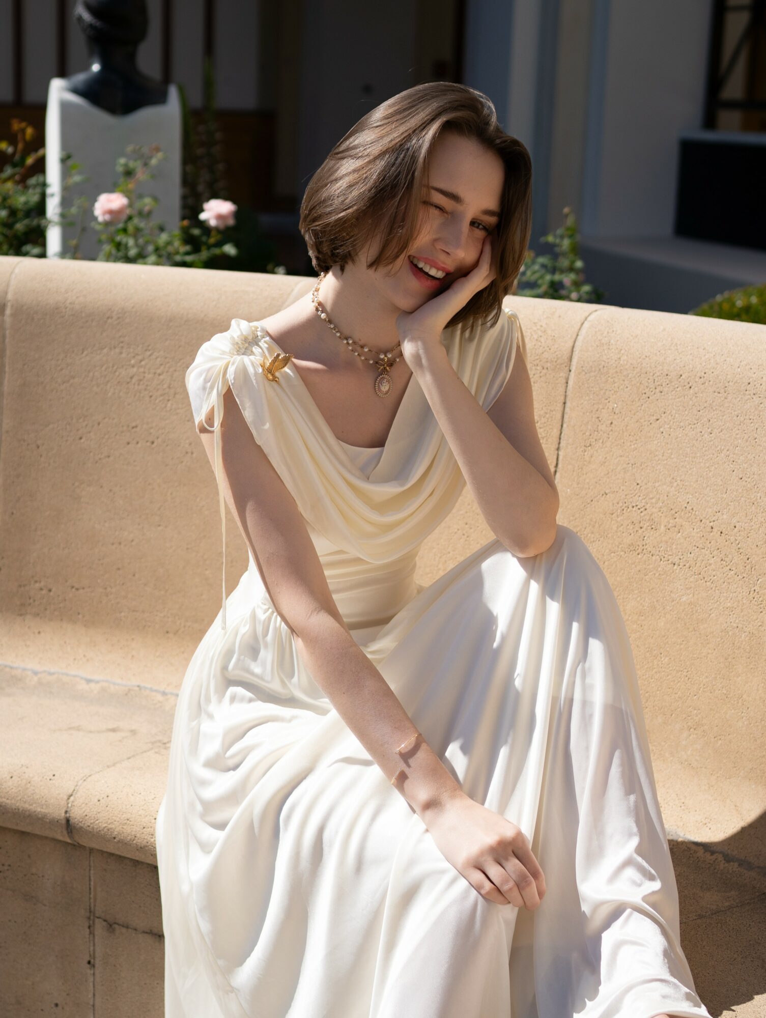Young woman with short hair sitting on a beige outdoor bench, smiling and laughing, wearing a white dress and necklace.