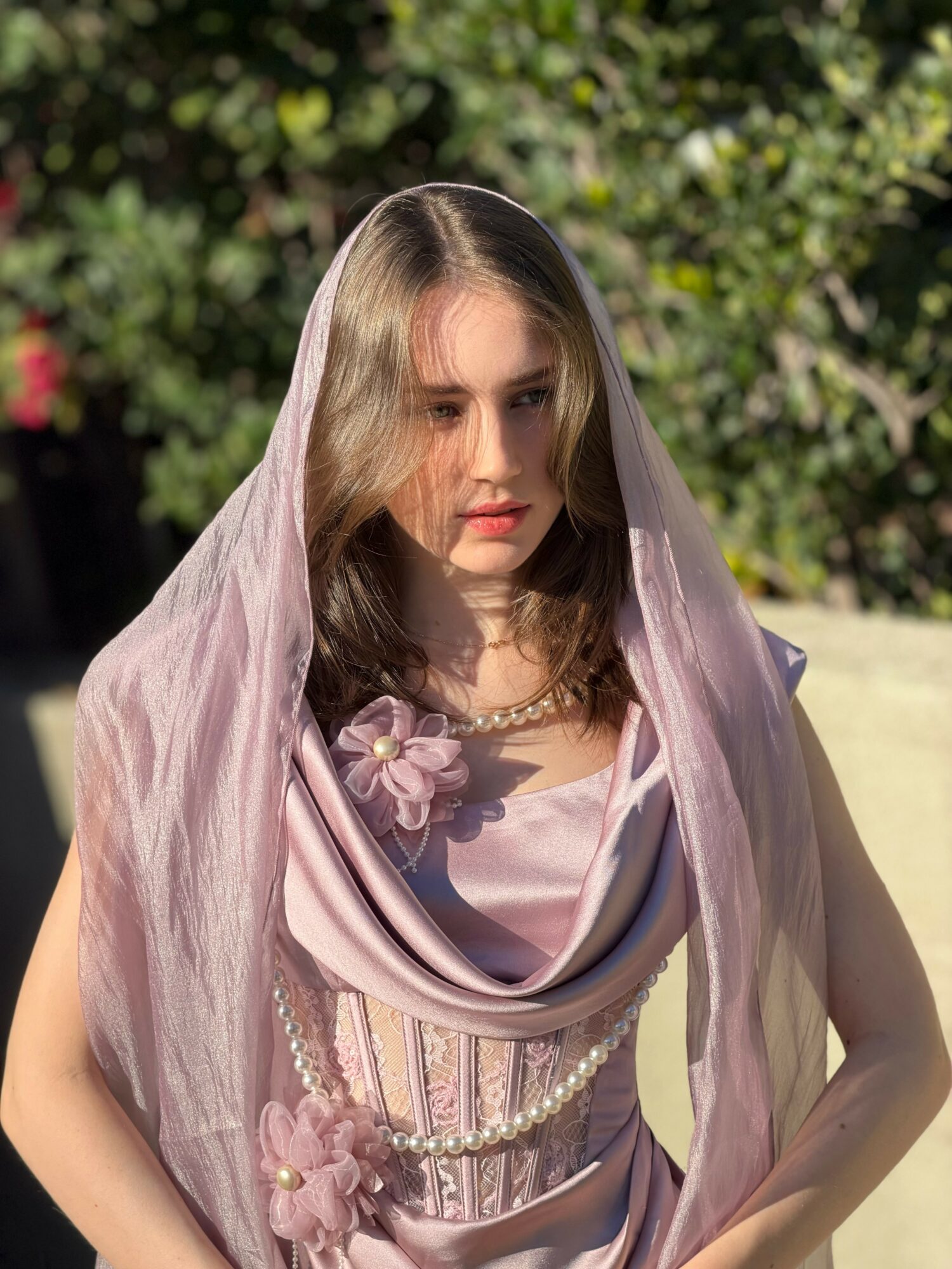 Young woman with long hair wearing a light pink dress and sheer veil outdoors, with greenery in background.
