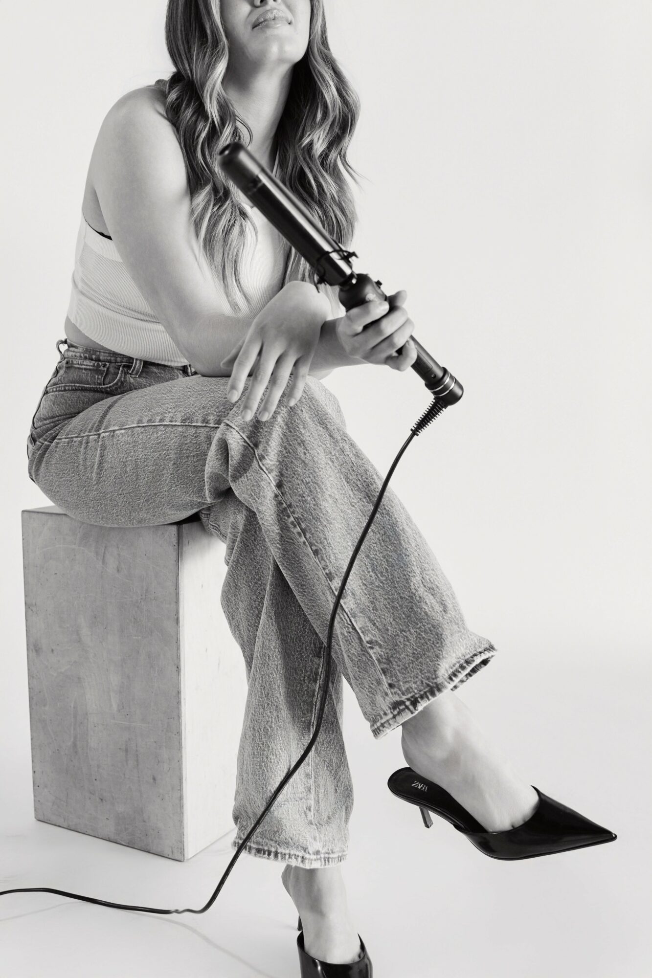 Woman sitting on a block holding a curling iron, wearing jeans and high heels, in black and white.