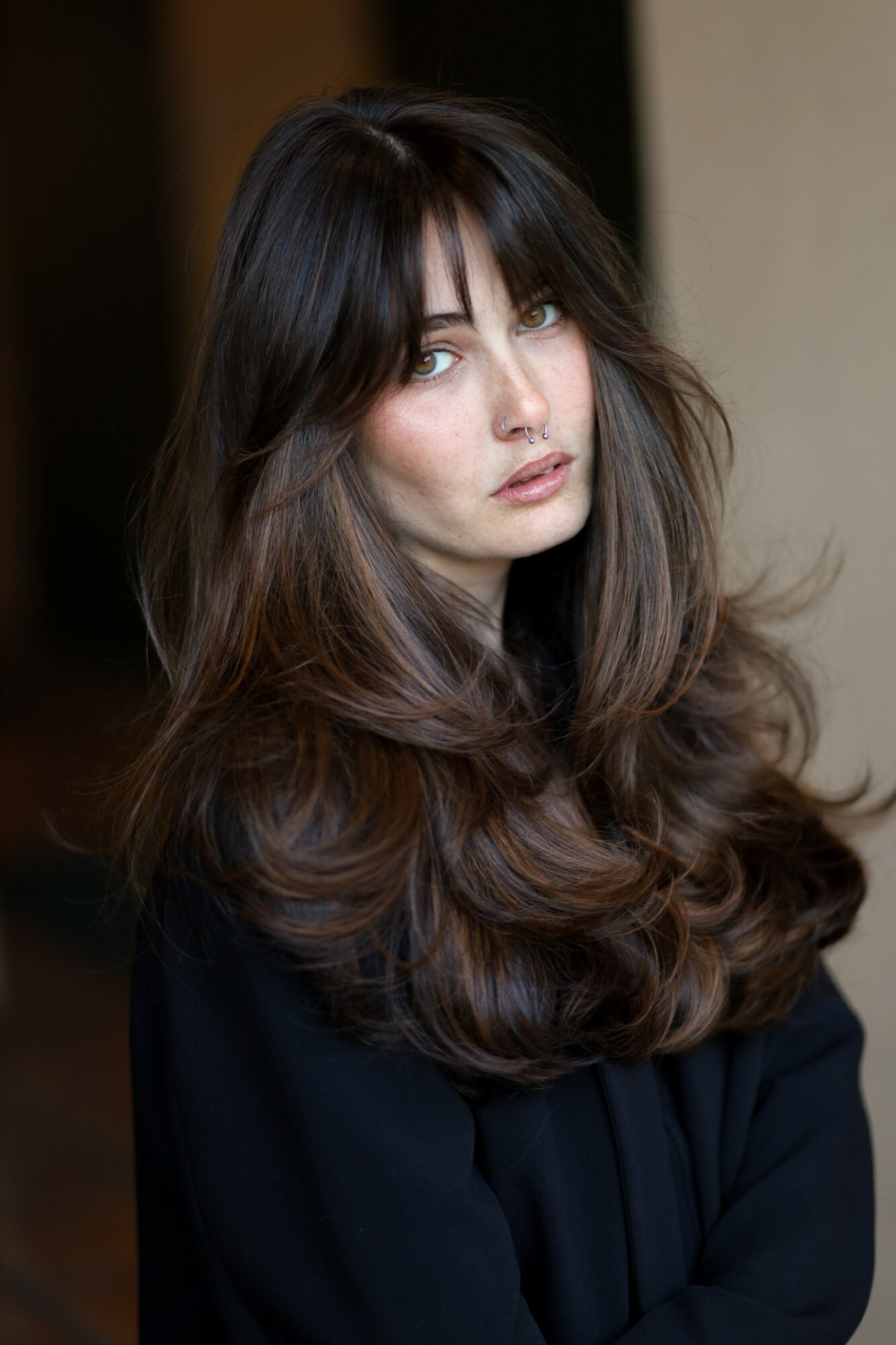 Young woman with long, wavy dark hair and bangs, looking at the camera, wearing a dark top, neutral background.