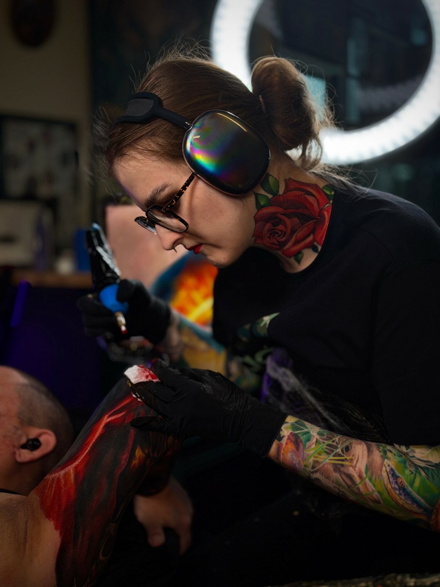 Tattooed person with glasses and large headphones tattooing a client in a tattoo studio, with a ring light in the background.