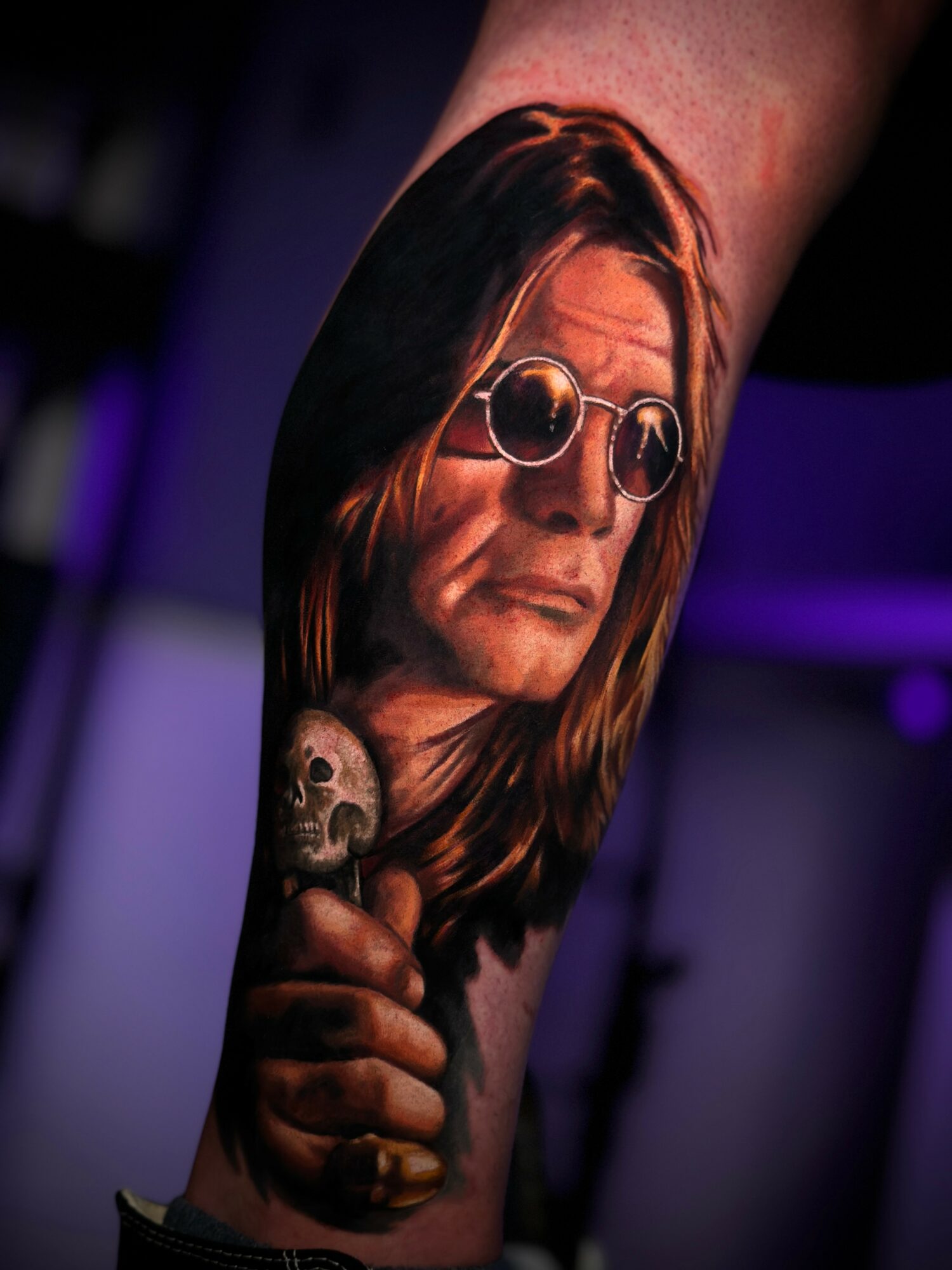 Tattoo of a man with long hair wearing sunglasses, holding a skull, with a dark background.