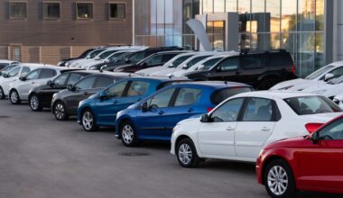 A row of various cars in a dealership parking lot, featuring blue, white, black, and red vehicles.
