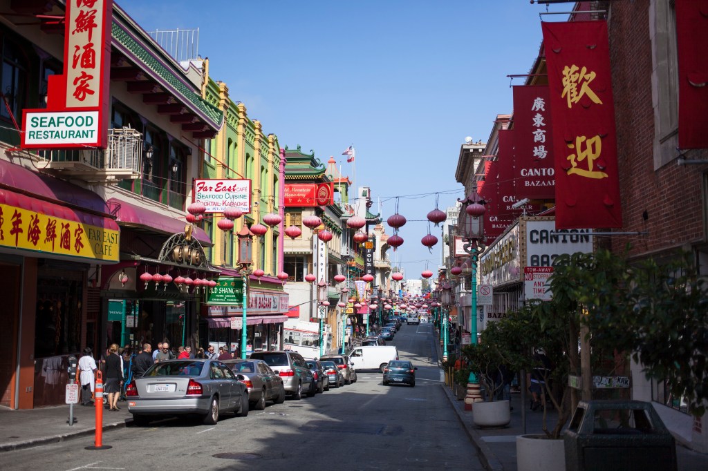 A car drives through Chinatown in San Francisco, with buildings displaying colorful signs and lanterns hanging overhead.