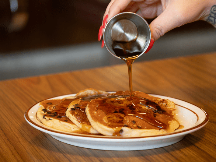 a woman's hand pours syrup on pancakes