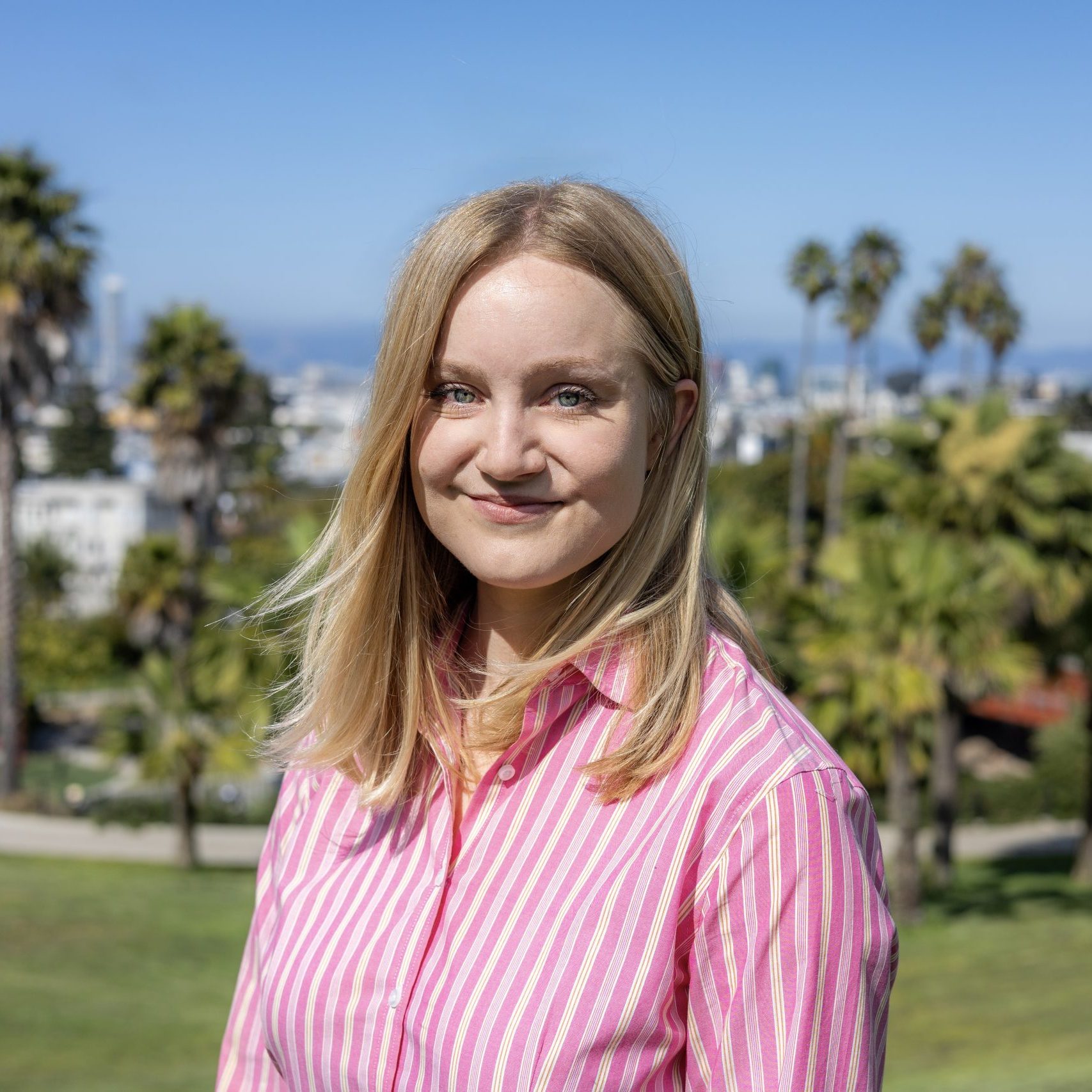 Woman with straight blonde hair and a pink striped shirt stands outside in a park with palm trees and a cityscape in the background on a sunny day.