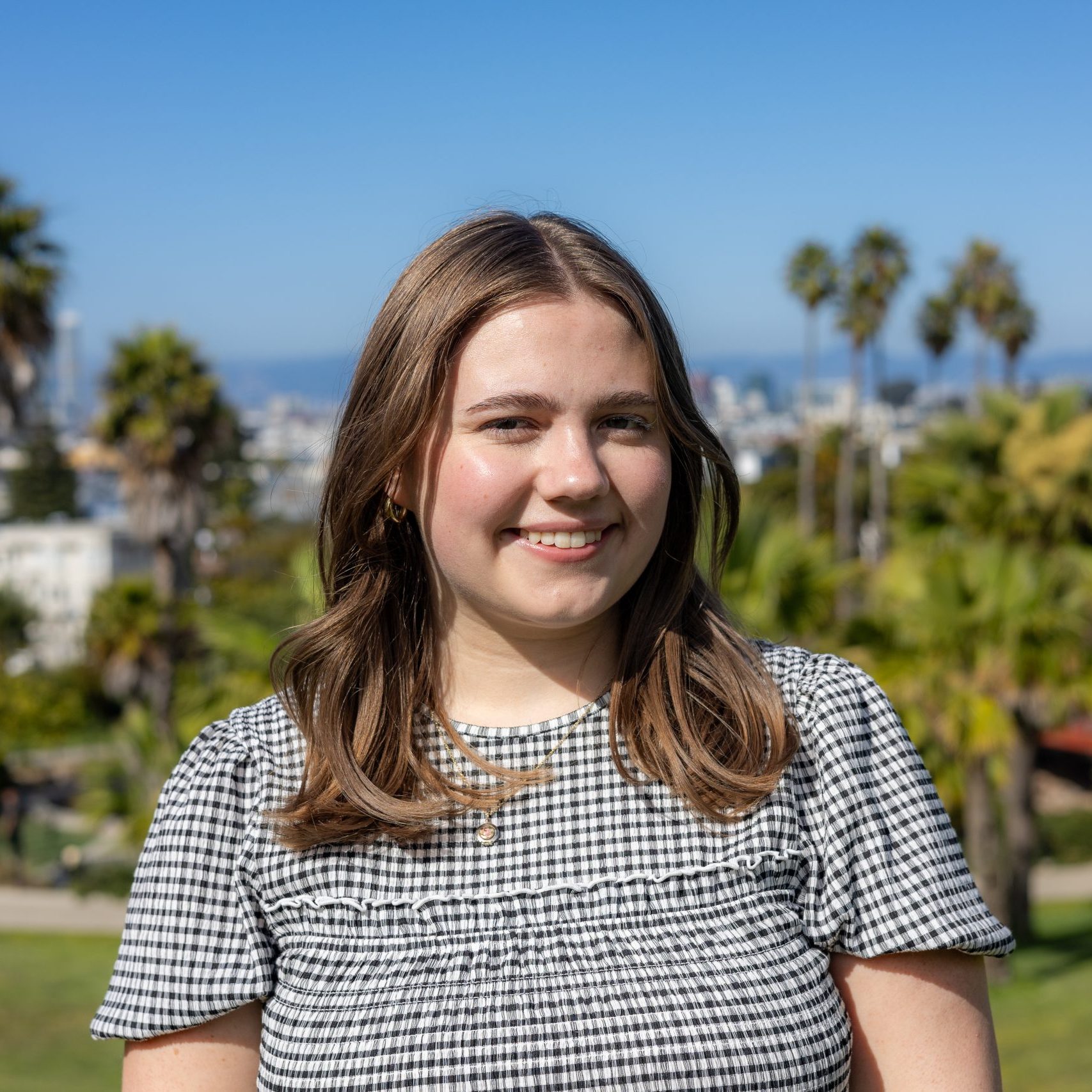 A young woman with shoulder-length brown hair, wearing a black and white checkered top, stands outside with palm trees and a clear blue sky in the background.