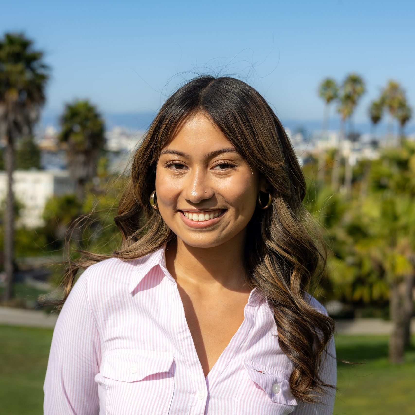 A woman with long, wavy hair and a pink striped shirt smiles outdoors in a park with palm trees and a cityscape in the background.