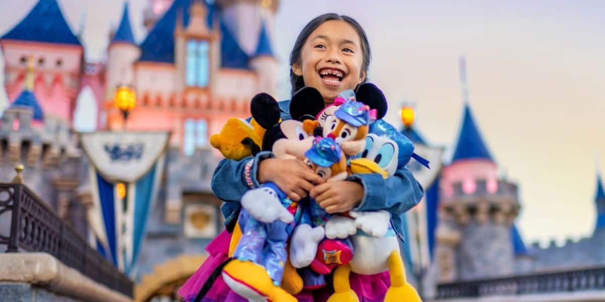 A joyful child holds several Disney character plush toys in front of a fairy-tale castle at Disneyland Resort. The background features turrets and decorations, enhancing the celebration atmosphere. The child is smiling widely, wearing a denim jacket.