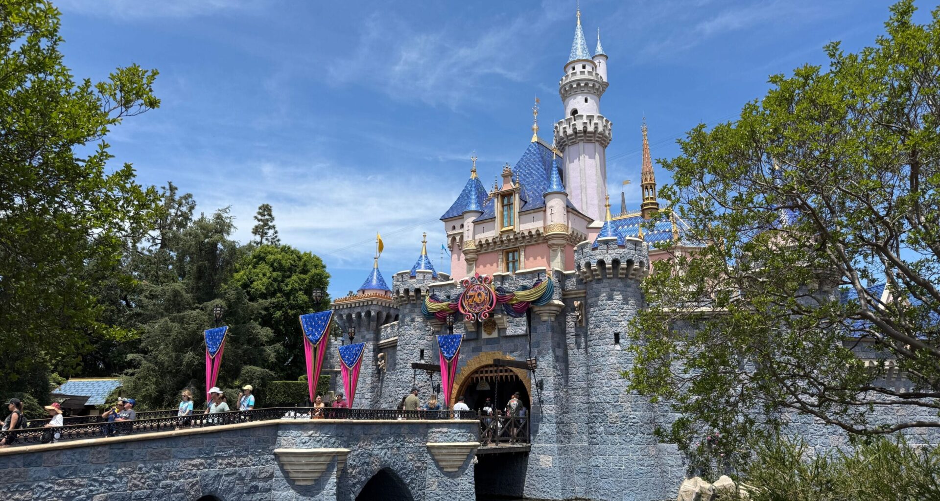 Sleeping Beauty Castle at Disneyland with blue sky, trees, and visitors crossing the stone bridge over a moat.