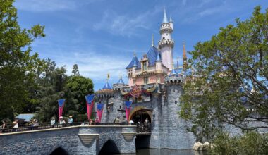 Sleeping Beauty Castle at Disneyland with blue sky, trees, and visitors crossing the stone bridge over a moat.